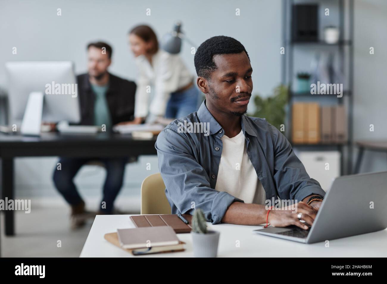 Afrikanischer junger Geschäftsmann in legerer Kleidung sitzt an seinem Arbeitsplatz und tippt im Büro auf dem Laptop Stockfoto