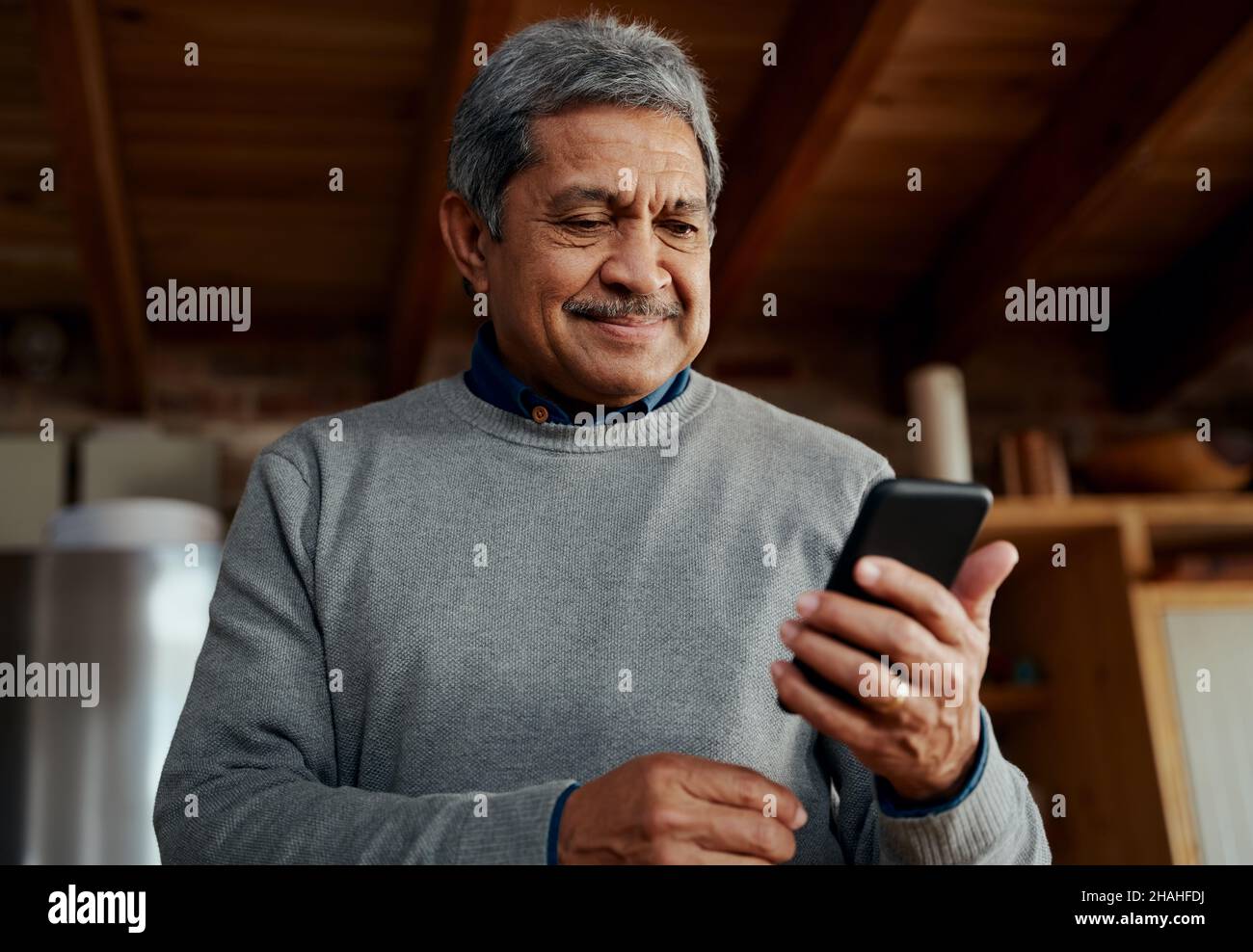 Lächelnder, multikultureller älterer Mann, der eine Nachricht auf dem Smartphone liest, während er in der modernen Küche steht. Stockfoto