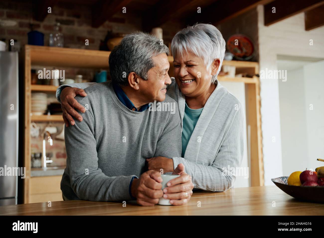 Glücklich pensionierte ältere Birazialpaare lächeln sich gegenseitig an. Frau hält Mann in der modernen Küche. Stockfoto