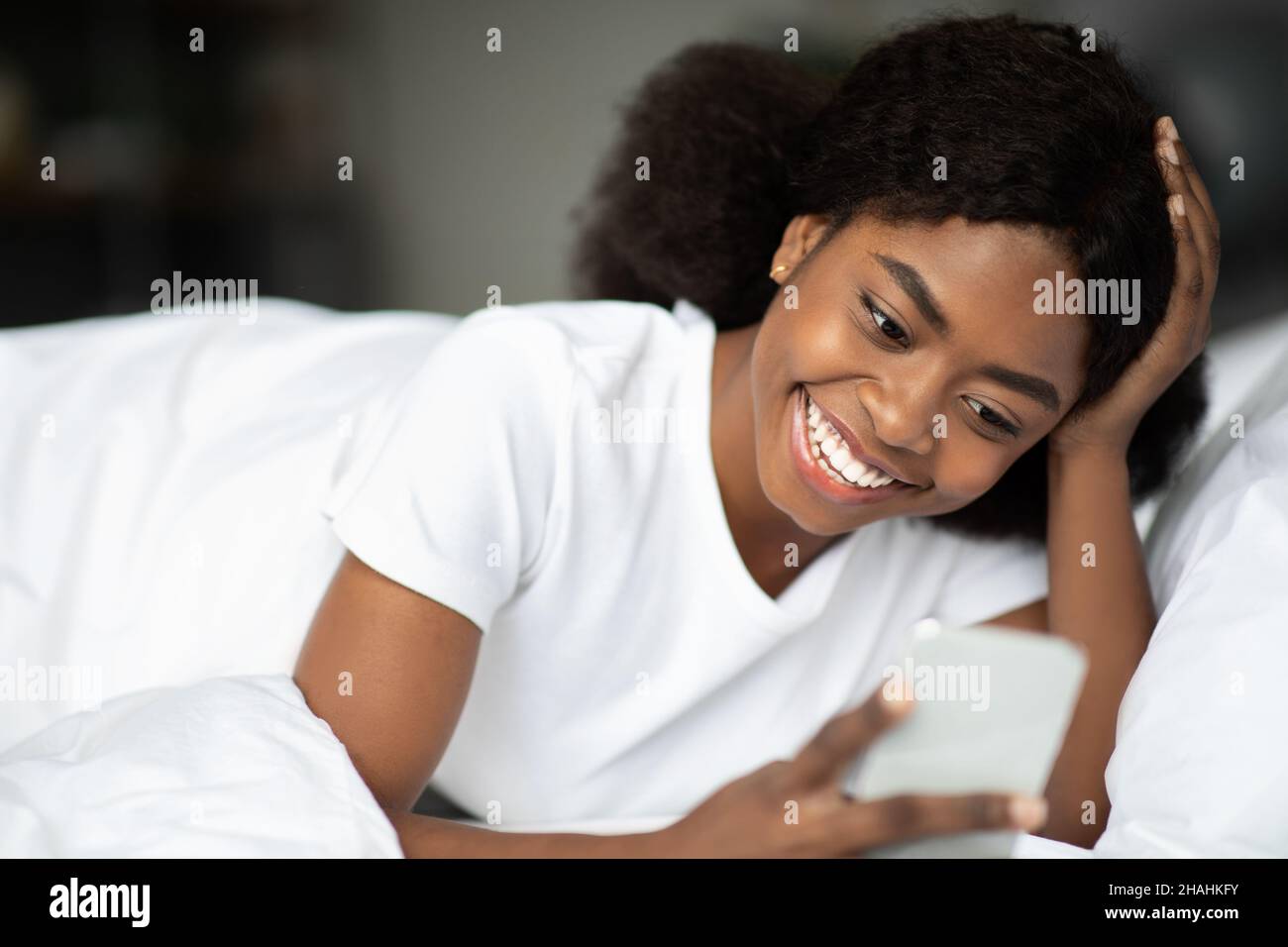 Glückliche afroamerikanische Frau, die im Bett liegt und SMS liest Stockfoto