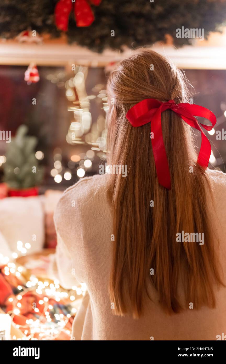 Gesichtslose Frau mit langen blondie Haaren und roten Bogen Haarclip Genuss zu Hause entspannen in gemütlichen bequemen Bett mit leuchtenden Lichtern Girlande Bokeh in der Nacht auf Stockfoto