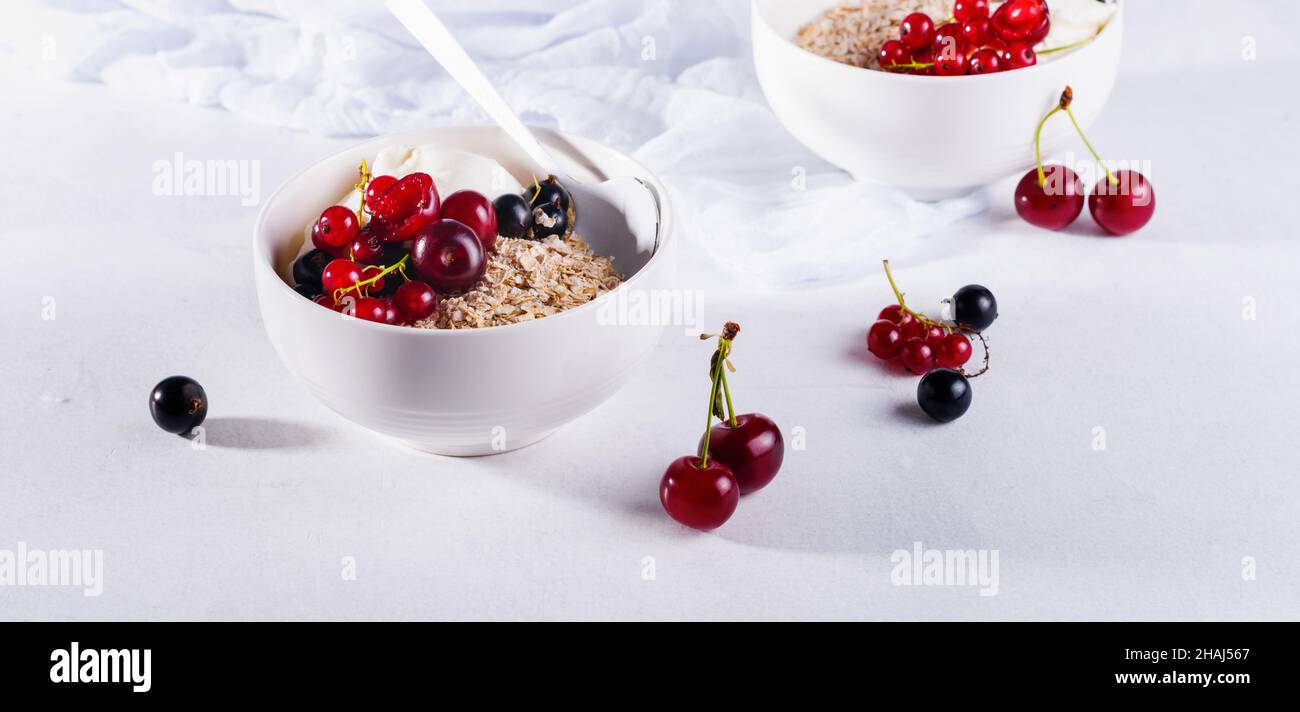 Roher Haferflocken mit Joghurt und Beeren aus Kirsche, Johannisbeere und Heidelbeere für ein gesundes Frühstück in einer weißen Keramikschale auf hellem Hintergrund. Selecti Stockfoto