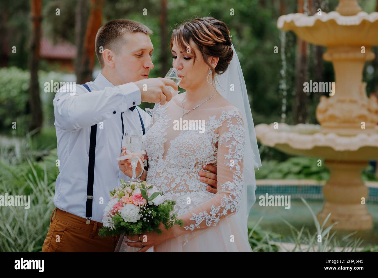 Braut und Bräutigam mit einem Glas Champagner im Park feiern ihren Hochzeitstag. Ein liebevolles Paar von Brautjungern trinkt Champagner, Umarmungen und Küsse Stockfoto