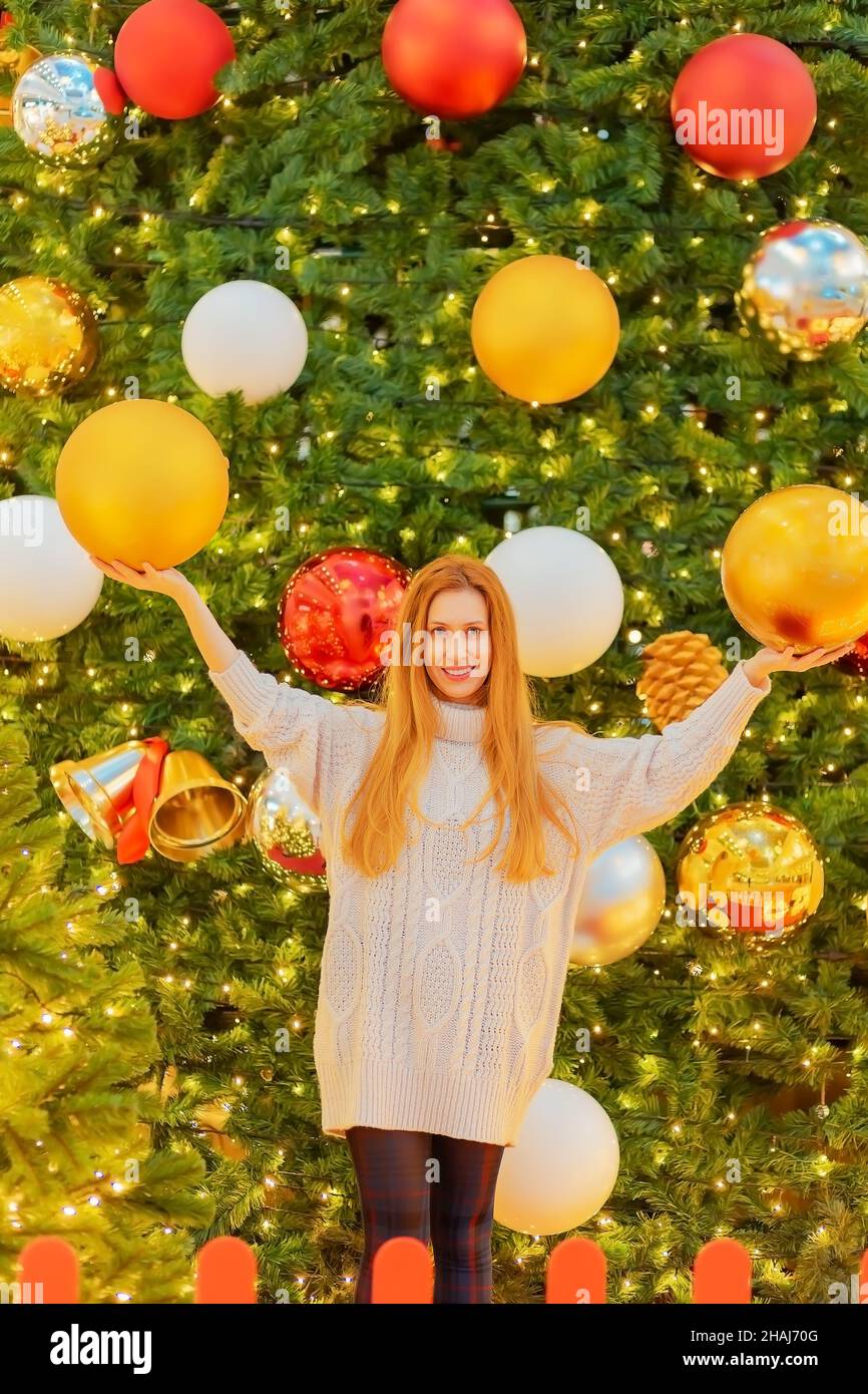 Lächelndes Mädchen hält riesige weihnachtskugeln, während sie in der Nähe des riesigen funkelnden weihnachtsbaums steht Stockfoto