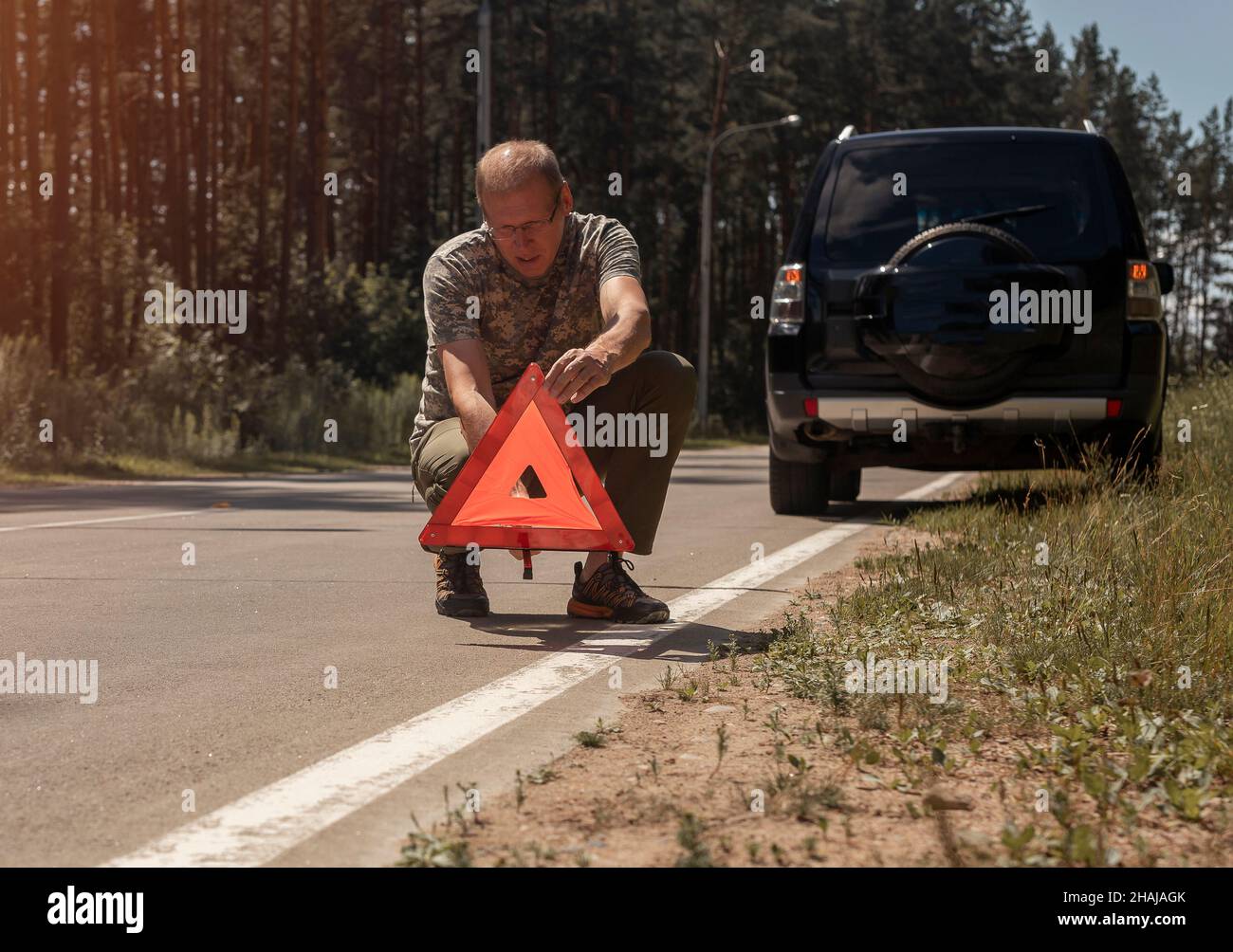 Reifer Mann Fahrer setzen Dreieck Vorsicht und Warnung Straßenschild auf Asphalt in der Nähe von Auto nach Unfall im Sommer. Stockfoto