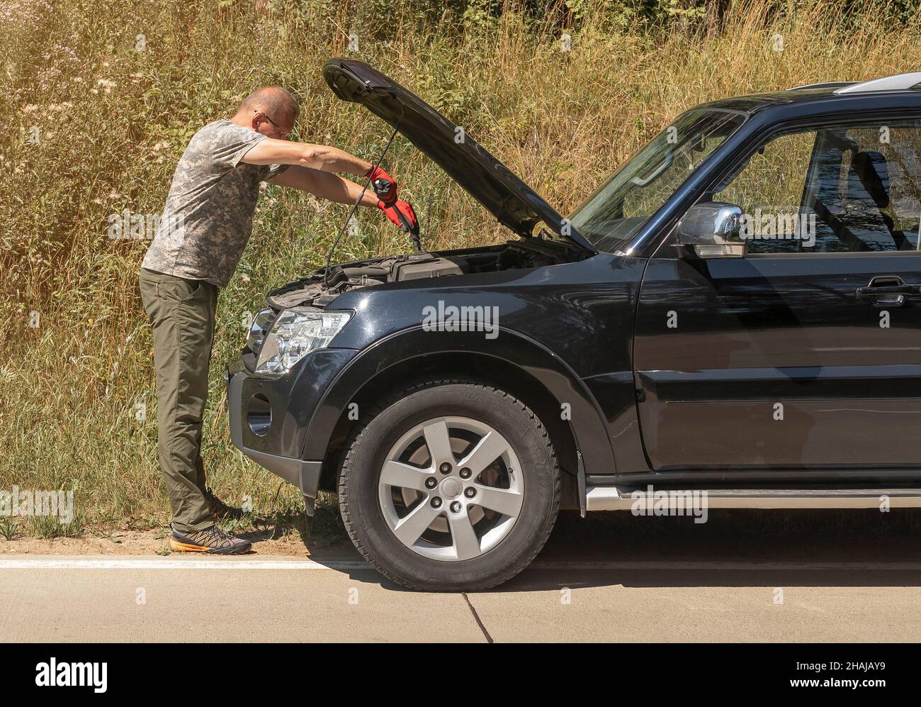 Mann, der im Sommer mit einem mechanischen Werkzeug in der Nähe eines Autos mit einer Haube im Freien steht und es nach der Autopanne auf der Reise repariert. Stockfoto