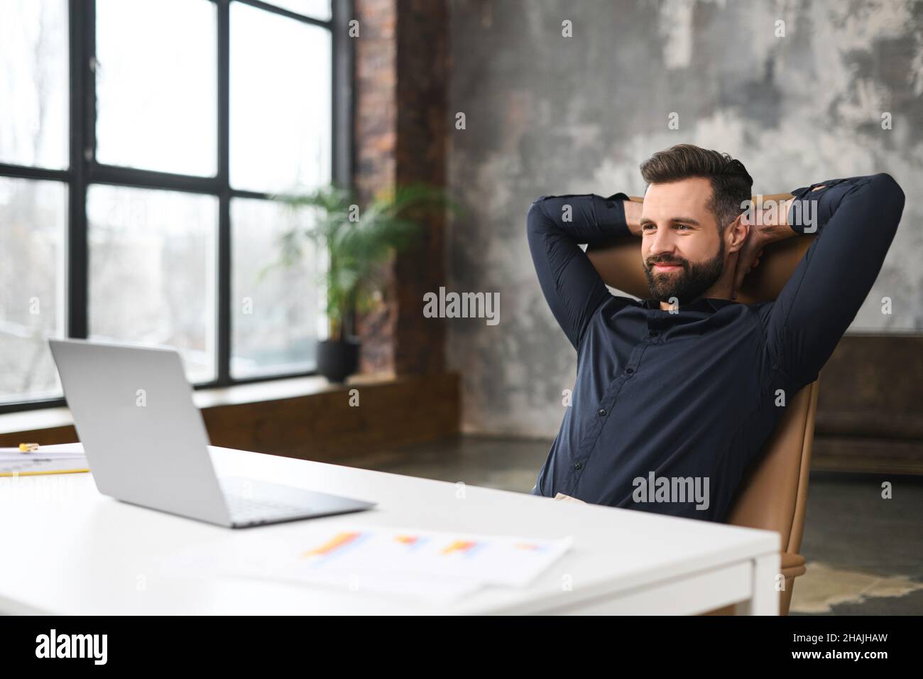 Unberasierter Mann, der am Tisch sitzt und lächelt, während er sich an seinem Arbeitsplatz wohlfühlt und entspannt. Selbstbewusster Büromitarbeiter, der vor dem Laptop sitzt Stockfoto