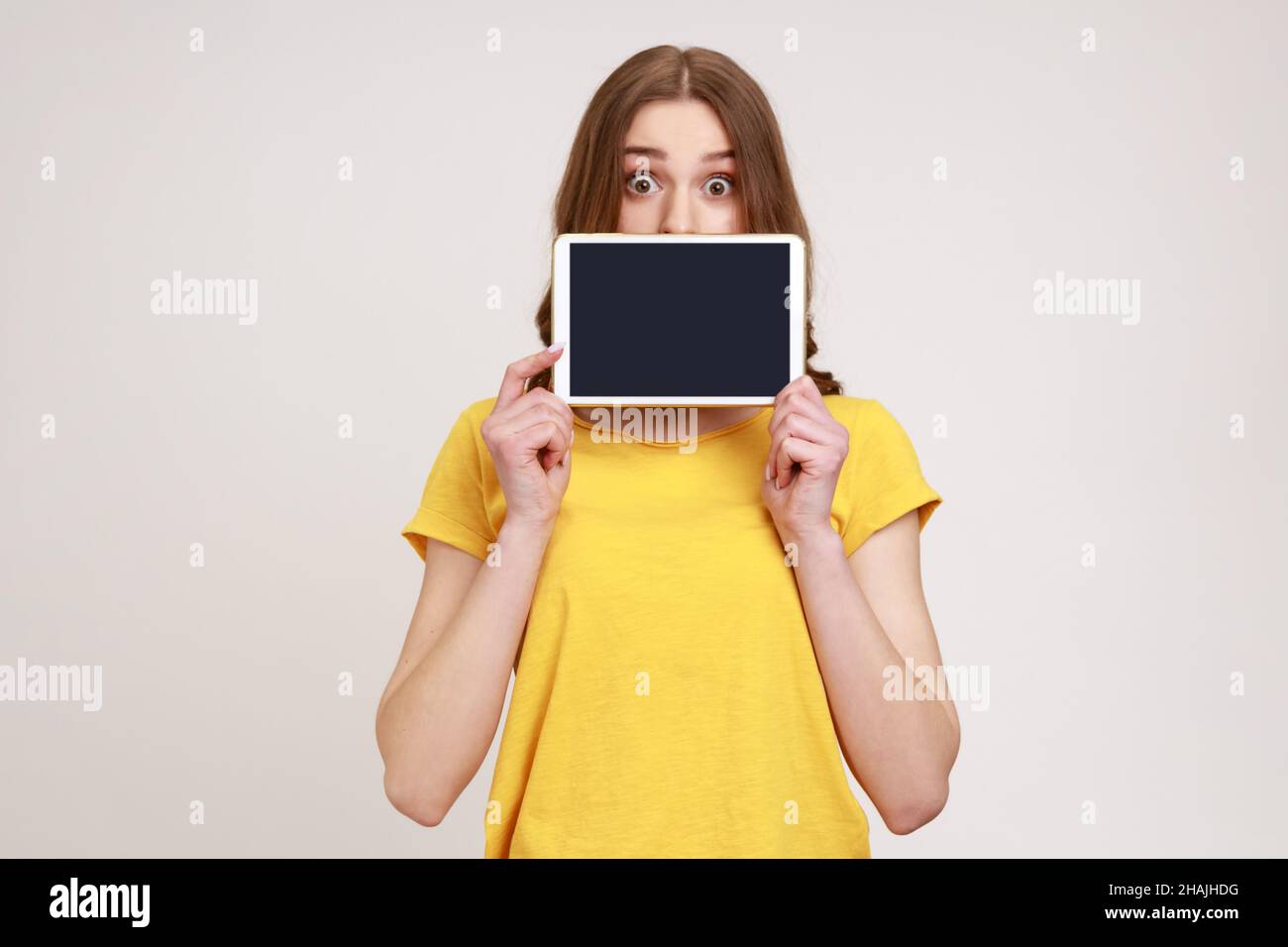 Porträt eines schönen Mädchen in gelben T-Shirt mit elektronischem Gerät, Blick auf die Kamera mit großen Augen, zeigt Tablet mit leeren schwarzen Bildschirm. Innenaufnahme des Studios isoliert auf grauem Hintergrund. Stockfoto