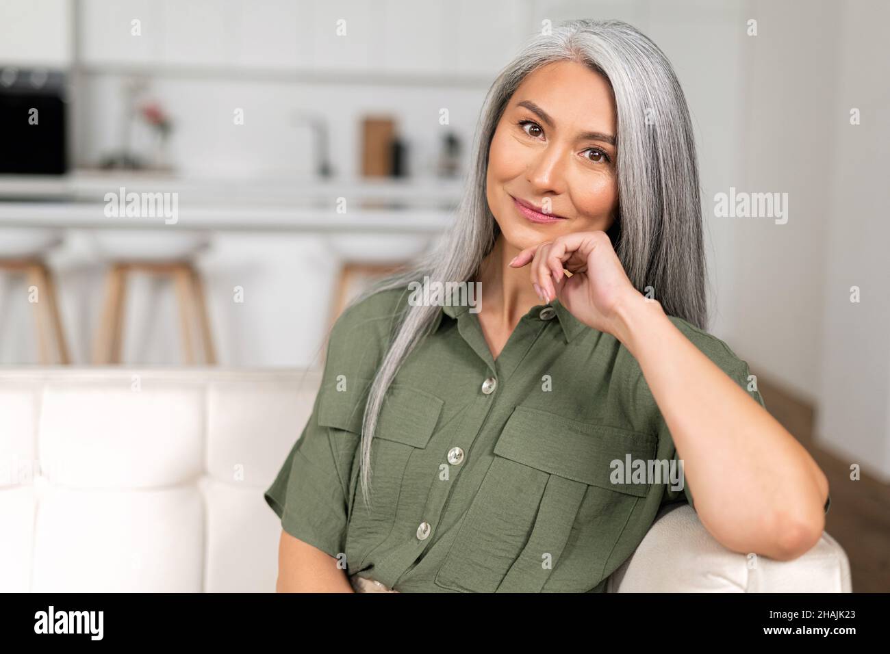 Nahaufnahme des Porträts einer charmanten Frau mittleren Alters mit langen silbernen Haaren, die in einer entspannten Atmosphäre sitzt, einer reifen asiatischen Frau aus dem Jahr 50s, die sich zu Hause ausruhte, die Kamera ansah und mit einem ruhigen Lächeln lächelte Stockfoto