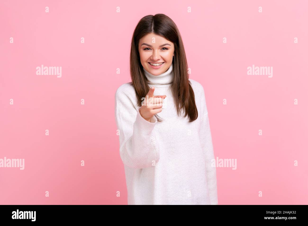 Glückliche positive Frau, die auf die Kamera zeigt, positive Emotionen zum Ausdruck bringt, Sie wählt, trägt weißen Pullover im lässigen Stil. Innenaufnahme des Studios isoliert auf rosa Hintergrund. Stockfoto