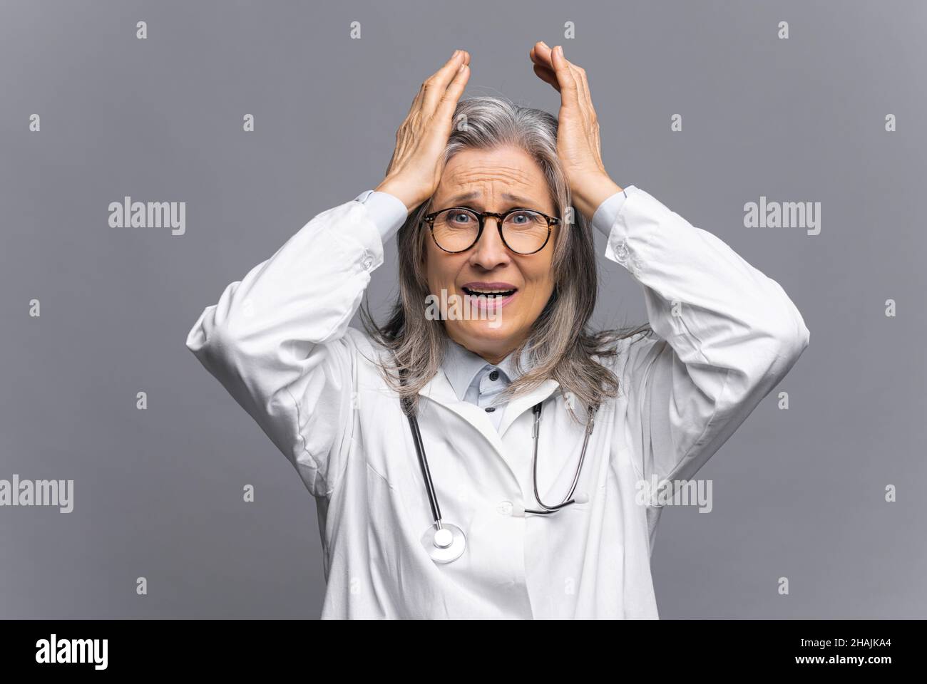 Leitender Arzt im weißen medizinischen Gewand mit Stethoskop auf den Schultern starrt mit Angst auf dem Gesicht auf die Kamera, greift den Kopf isoliert auf grau, reifer Physiotherapeut fühlt Verzweiflung und Verzweiflung Stockfoto