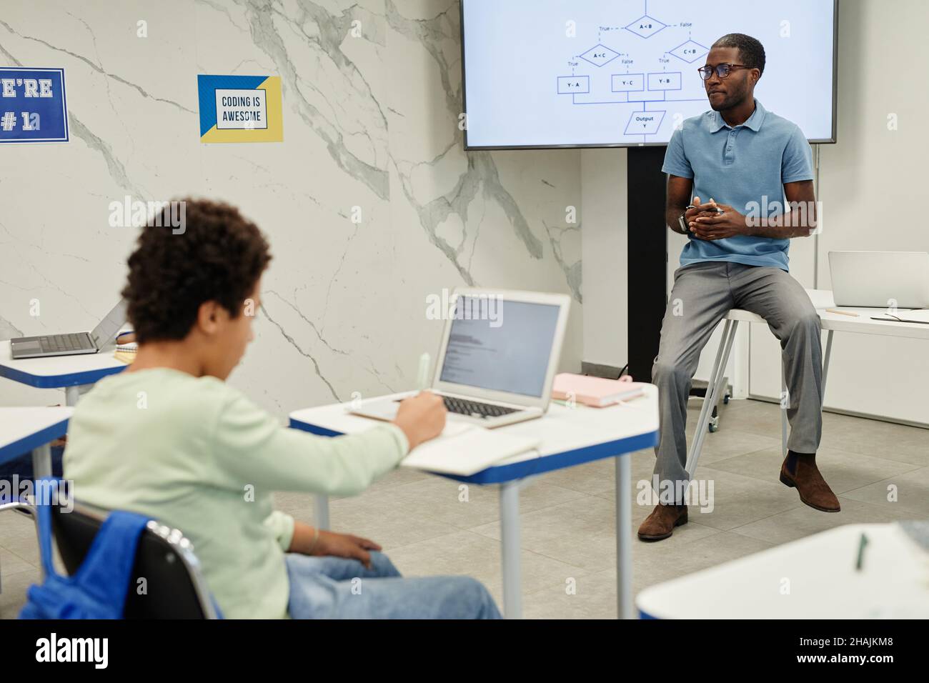 Porträt eines jungen afroamerikanischen Lehrers, der auf dem Schreibtisch sitzt, während er Codierungsunterricht für Kinder unterrichtet, Kopierraum Stockfoto