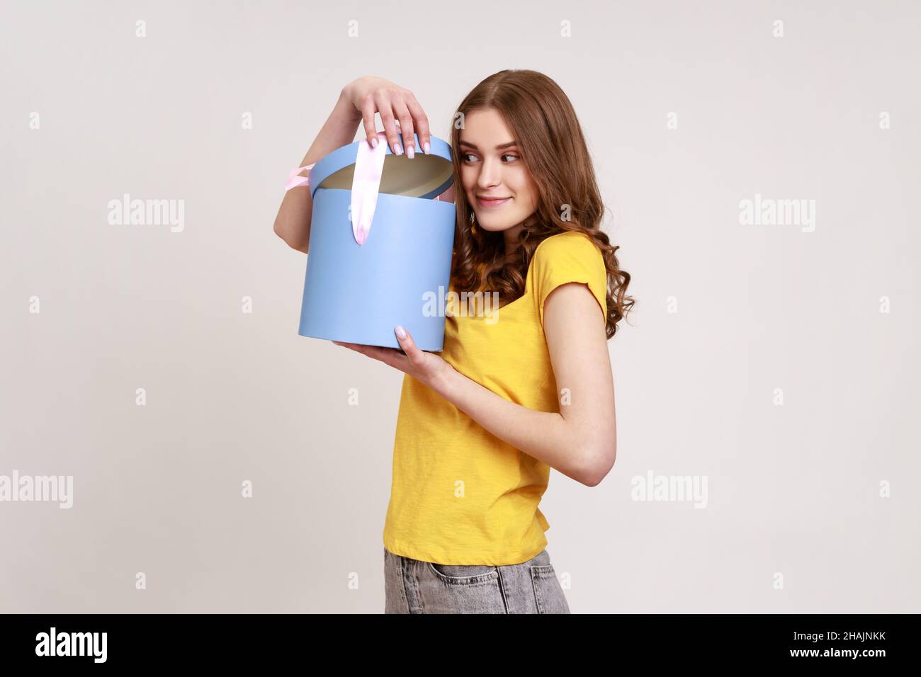 Porträt von neugierigen glücklich braun behaart Teenager-Mädchen in gelben T-Shirt, Eröffnung Geburtstagsgeschenk, Blick auf die Kamera mit Interesse, Auspacken Geschenk. Innenaufnahme des Studios isoliert auf grauem Hintergrund. Stockfoto