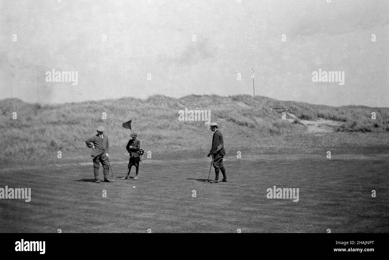 Um 1910, historisch, auf einem Links-Golfplatz, auf einem Putting Green, zwei Golfer in der formellen Kleidung der Ära, einer der in plus zwei, beide mit Jacken, mit einem jungen Caddy, der die Flagge hält, England, Großbritannien. Stockfoto