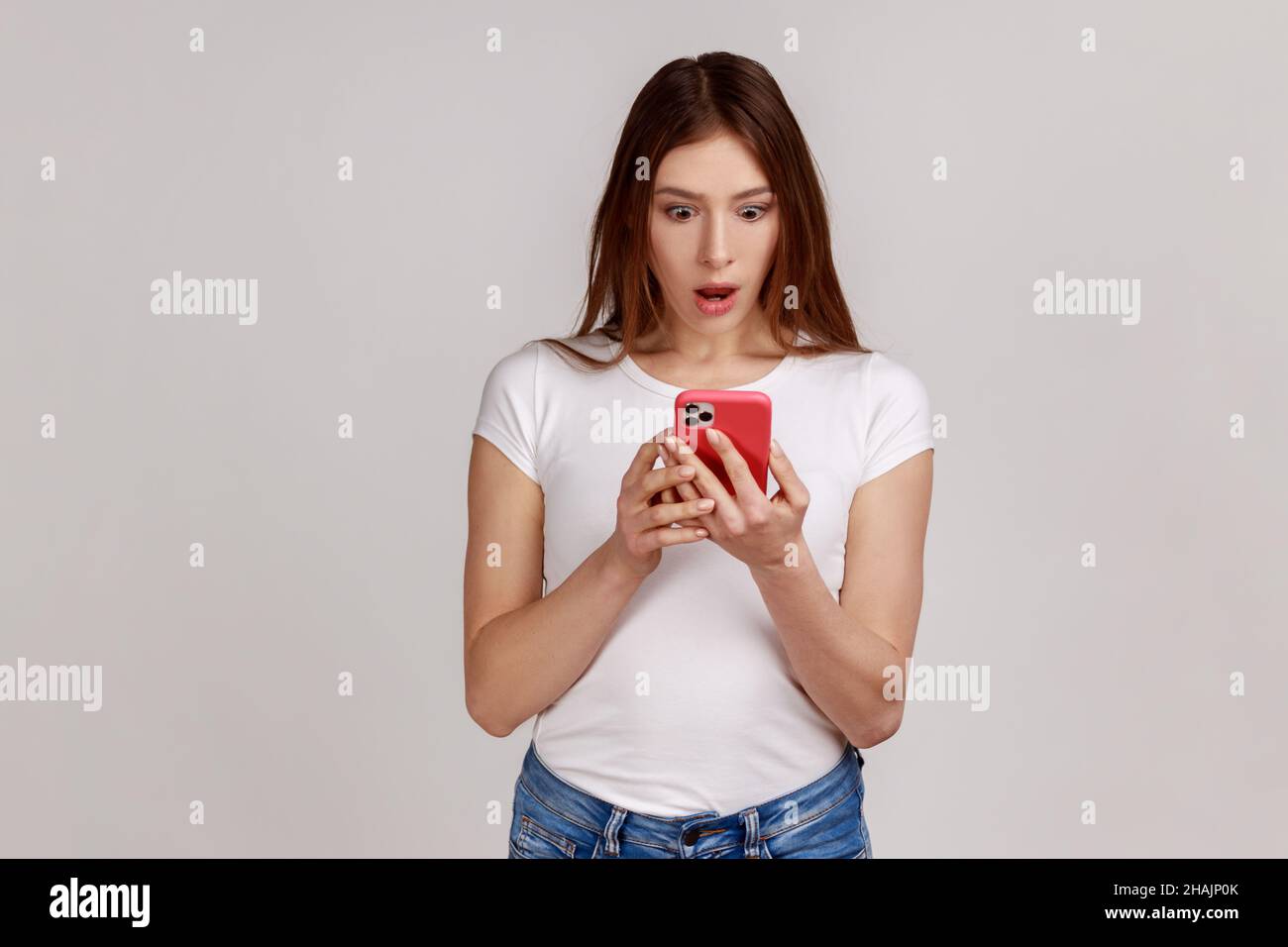 Porträt einer schockierten, erstaunten Frau, die per Handy einen Beitrag in einem sozialen Netzwerk liest, chattet, überrascht aussieht und ein weißes T-Shirt trägt. Innenaufnahme des Studios isoliert auf grauem Hintergrund. Stockfoto