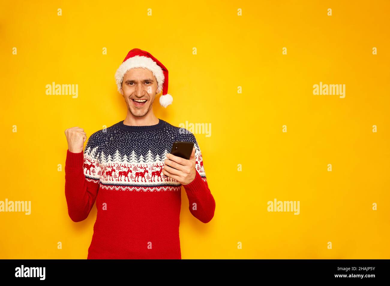 Aufgeregt Mann in Neujahr Pullover, roten Weihnachtshut, isoliert gelben Hintergrund mit freiem Platz für Text der Werbung, mit Telefon in den Händen zeigt Siegeszeichen. Konzept - Gewinn, Verkauf, Rabatte. Hochwertige Fotos Stockfoto