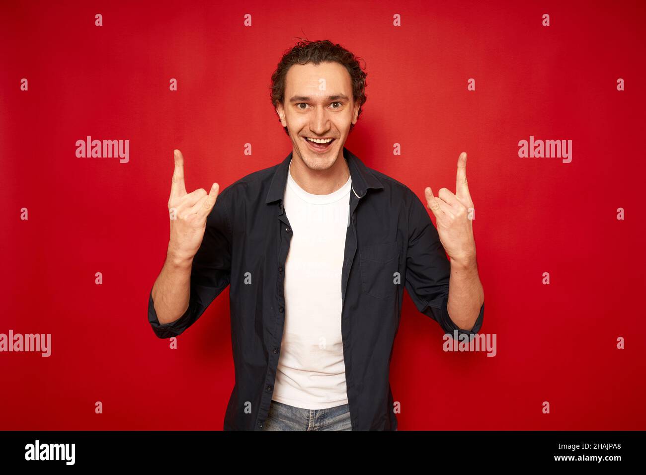 Glücklich überglücklich schöner Mann mit lockigen Haaren, zeigt coole Zeichen Liebe, sagt: Ja, dieser wunderbare hübsche junge Mann Gesten innen vor rotem Hintergrund Kopierraum. Menschen und Körpersprache Konzept. Hochwertige Fotos Stockfoto