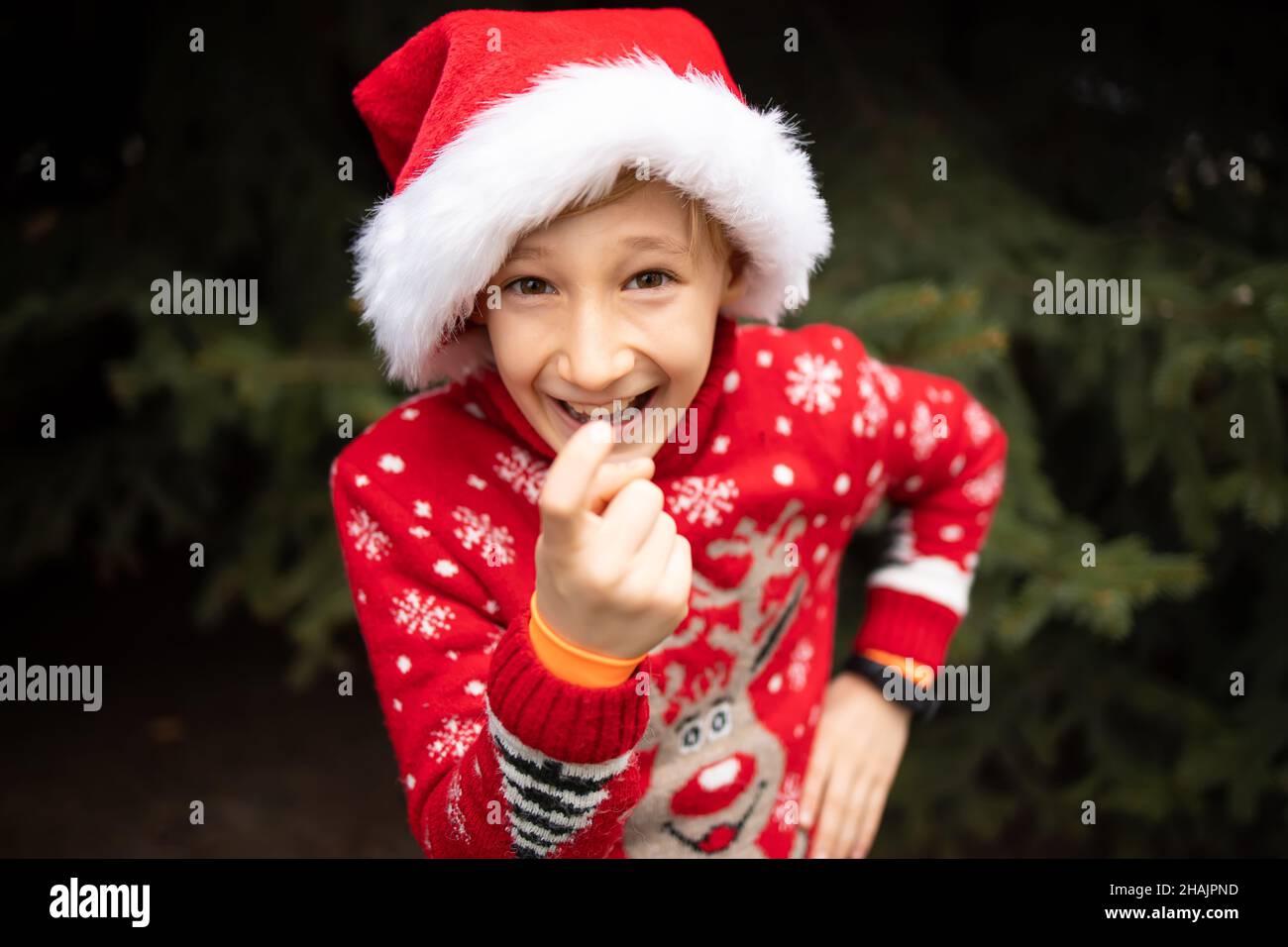 Ein Junge in einem rot gestrickten Weihnachtspullover mit einem Weihnachtsrentier und einem Weihnachtsmannhut lockt sich mit seinem Zeigefinger listig an Stockfoto