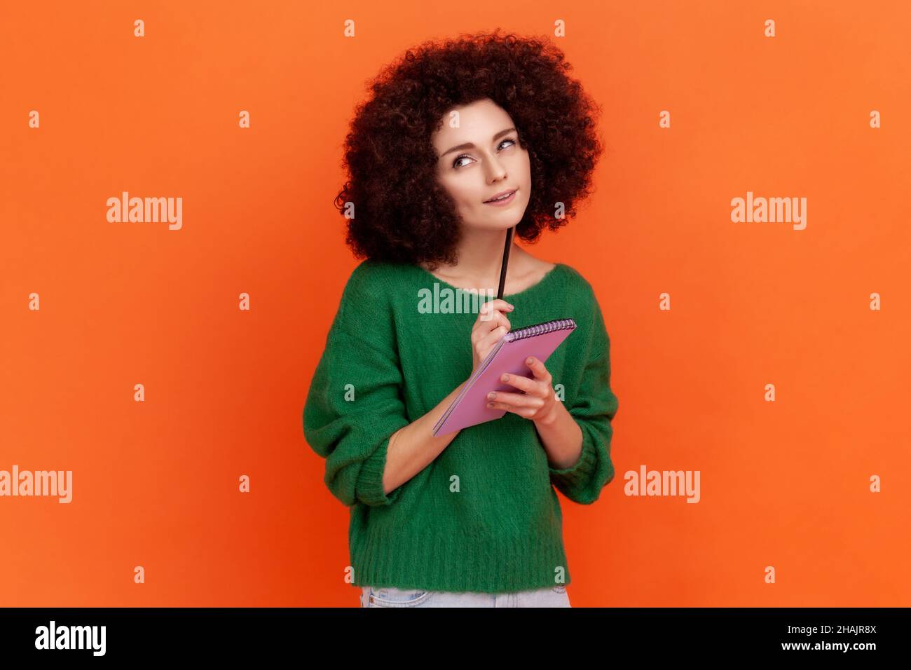 Gut aussehende nachdenkliche Frau mit Afro-Frisur trägt grünen lässigen Stil Pullover stehen mit Papier-Notizbuch und Stift, so dass zu tun Liste, Planung. Innenaufnahme des Studios isoliert auf orangefarbenem Hintergrund Stockfoto