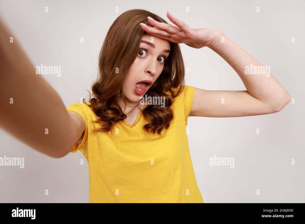 Erstauntes Teenager-Mädchen in gelbem legerem T-Shirt, das Selfie macht, auf den Blickwinkel der Kamera schaut, auf das Foto, weit über den Kopf schaut. Innenaufnahme des Studios isoliert auf grauem Hintergrund. Stockfoto
