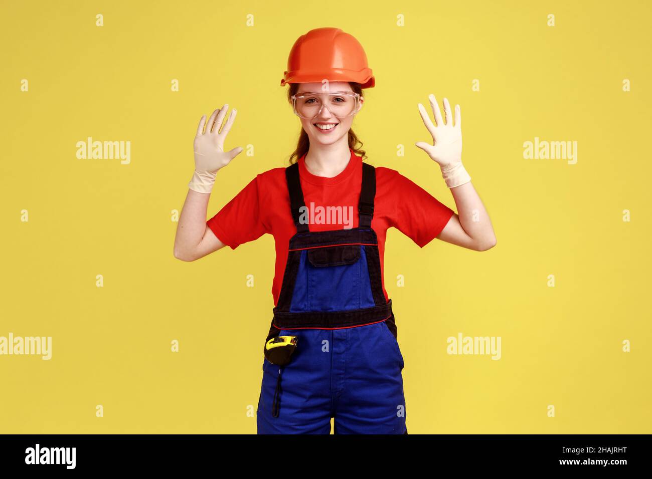 Positiv lächelnde Baumeisterin, die mit erhobenen Armen steht und ihre Gummihandschuhe zum Schutz der Hände zeigt, Overalls und Schutzhelm trägt. Innenaufnahme des Studios isoliert auf gelbem Hintergrund. Stockfoto