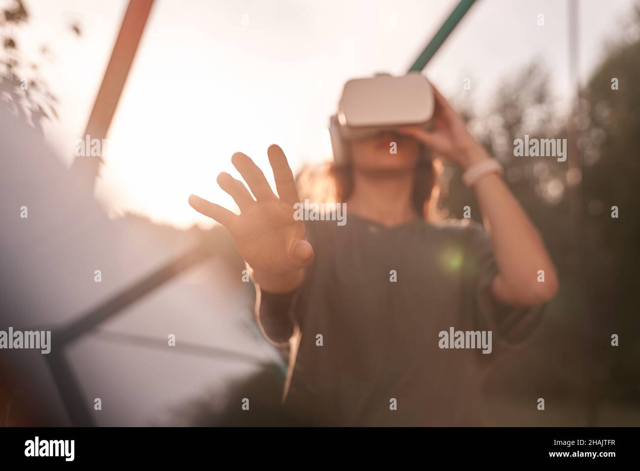 Nahaufnahme eines Mädchens, das mit den Händen eine Virtual-Reality-Brille auf dem Kopf hält und auf einem Hintergrund von grünen Bäumen steht. Er reicht eine Hand an die Kamera. Hochwertige Fotos Stockfoto