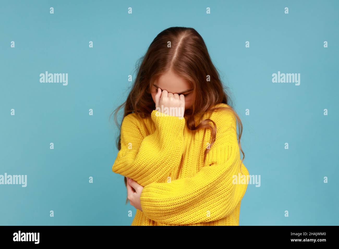 Porträt eines kleinen Mädchens versteckt Gesicht nach unten und weinen, aufgeregt über Verlust, Gefühl Trauer und Bedauern, tragen gelben lässigen Stil Pullover. Innenaufnahme des Studios isoliert auf blauem Hintergrund. Stockfoto