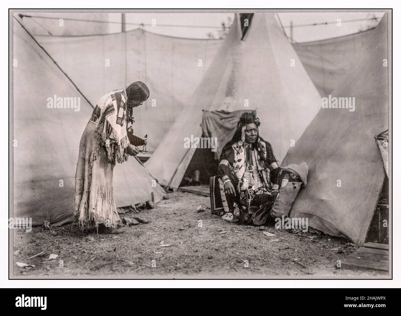Papoose nordamerikanische indianische Frau aus der Region des Nordwestplateaus, die mit einem Kodak-Filmbälge eine handgehaltene Kamera von einem Mann und einem Baby in einem Gepäckträger vor einem Tipis fotografiert. Die Leute sind möglicherweise Darsteller in einer Wild West Show oder einer Side Show. 1911 - Indianer Nordamerikas--Kleidung & Kleidung--Nordwesten, Pazifik--1910–1920 Kameras--1910–1920 Tipis--1910–1920 Silbergelatine-Drucke--1910-1920. Portraitfotos--1910-1920. Stockfoto