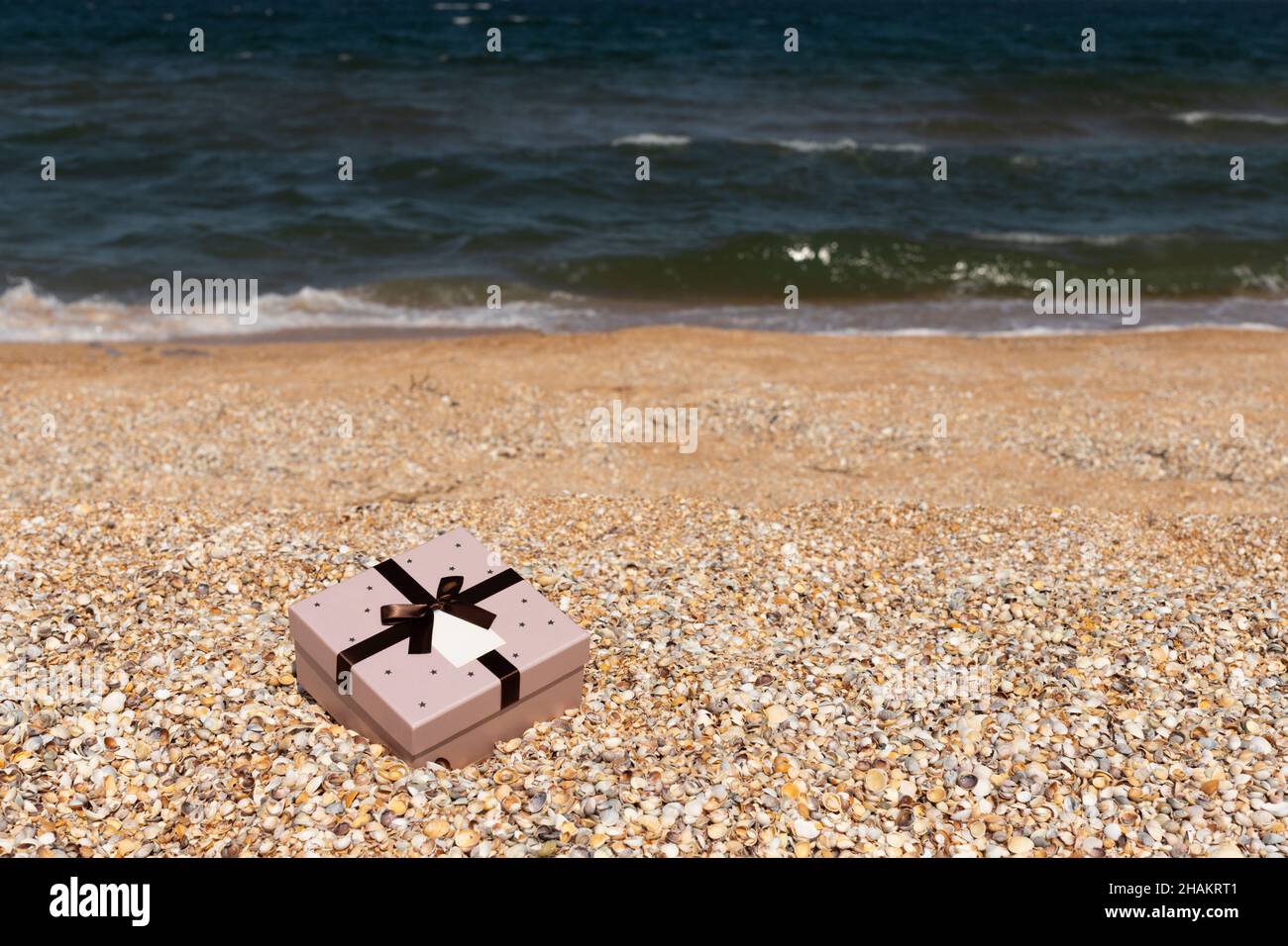 Am Strand in der Nähe des Meeres liegt eine Schachtel mit einem mit einem Band gebundenen Geschenk. Vorderansicht, vertikal. Stockfoto