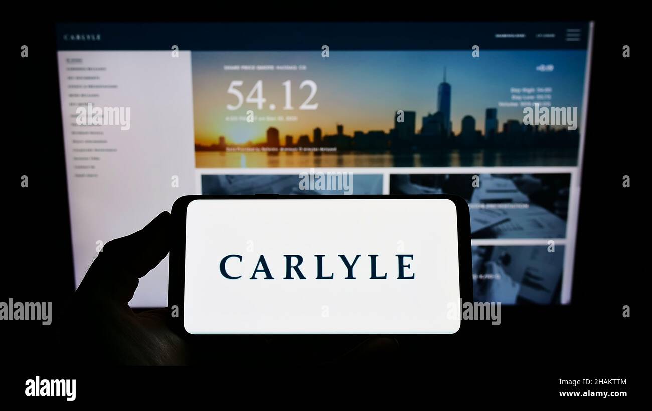 Person, die ein Mobiltelefon mit dem Logo der amerikanischen Investmentgesellschaft The Carlyle Group L.P. auf dem Bildschirm vor der Webseite hält. Konzentrieren Sie sich auf die Telefonanzeige. Stockfoto