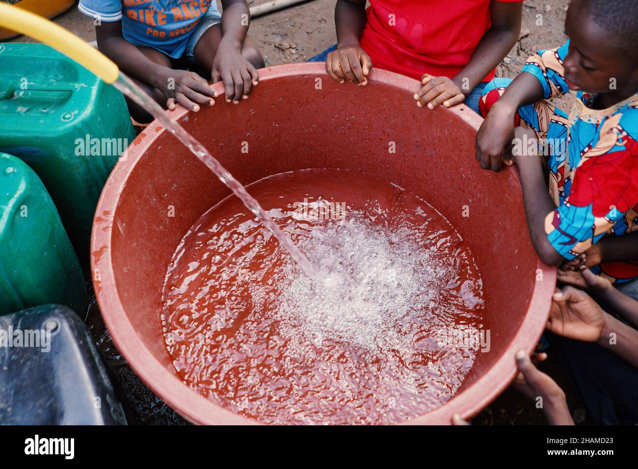 Nahaufnahme frischer, sauberer Wasserstrahl, der in den roten Eimer fließt und von durstigen afrikanischen Kindern umgeben ist, die warten Stockfoto