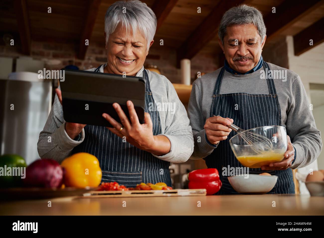 Lachende multikulturelle ältere Frau mit Tablet, um Rezept in der modernen Küche mit Ehemann zu erforschen. Glückliches Paar, das gesunde Mahlzeiten kocht. Stockfoto