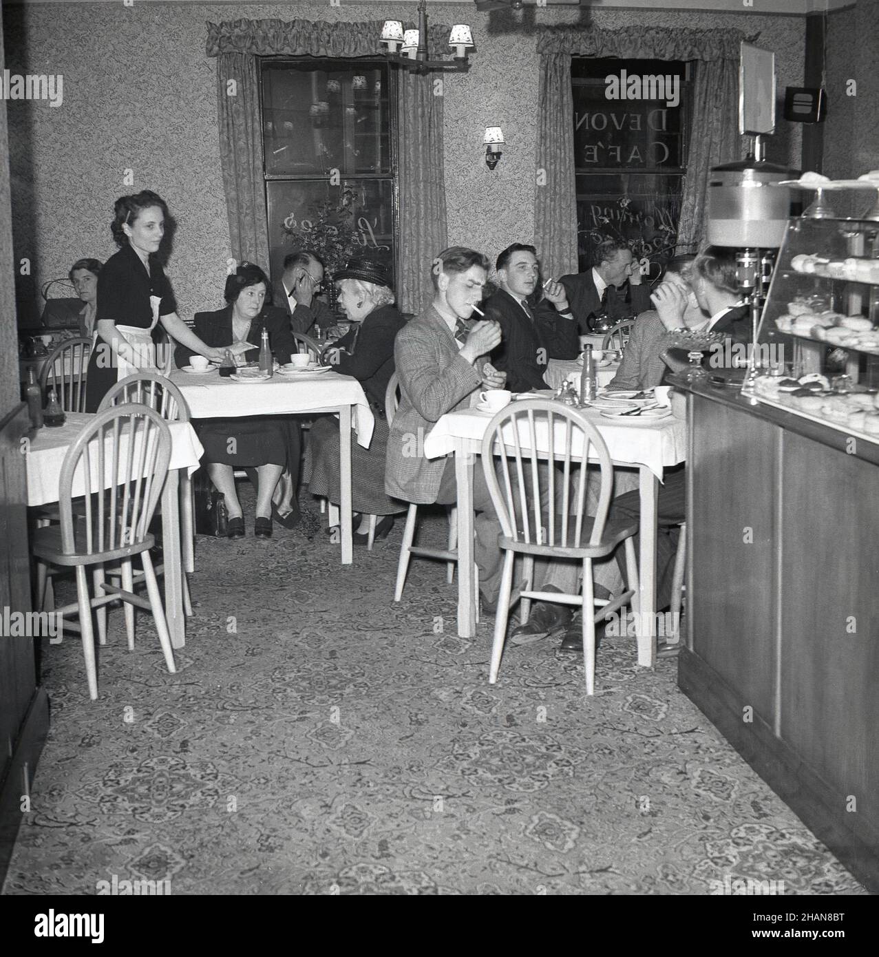 1950s, historisch, Kunden im oberen Essbereich eines Cafés der Ära, sitzen an mit Stoff bedeckten Tischen, Bedienung und mit einem Zähler mit Kuchen im Angebot, UK.das Fruchtgetränk, Qoush ist neben dem Schalter erhältlich. Quosh war ein aromatisierter Squash oder schwacher Cordial, dem Wasser hinzugefügt wurde und wurde erstmals im Vereinigten Königreich im Jahr 1950s in den Geschmacksrichtungen Orange, Gerstenwasser und Zitrone hergestellt. Stockfoto