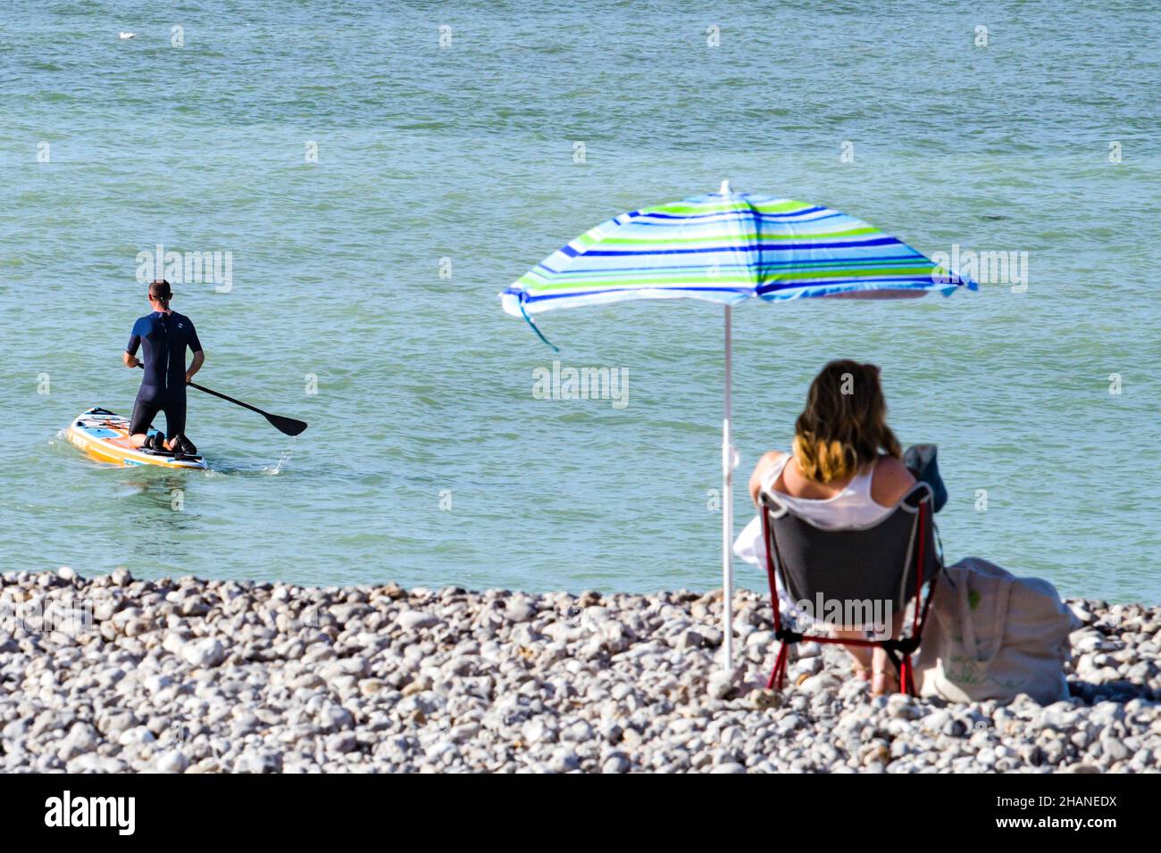 Saint-Valery-en-Caux (Nordfrankreich): Paddelboard und Frau, die unter einem Sonnenschirm am Kiesstrand sitzt Stockfoto