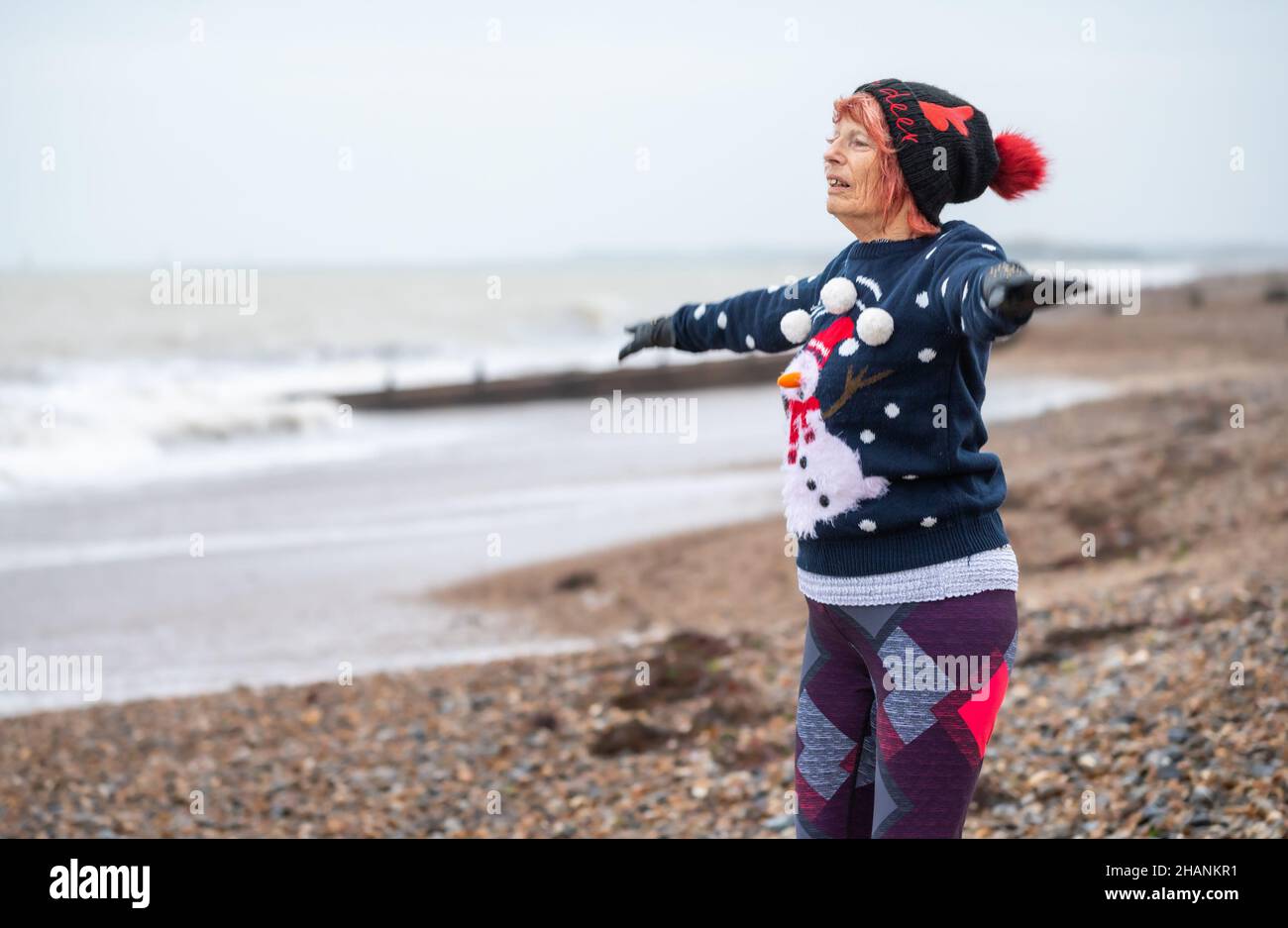 Ältere ältere Frau mit 80s Jahren an einem Strand, die im Winter trainiert. Sie trägt einen weihnachtlichen Pullover und Hut, während sie an der Südküste, Großbritannien, Sternsprünge macht Stockfoto