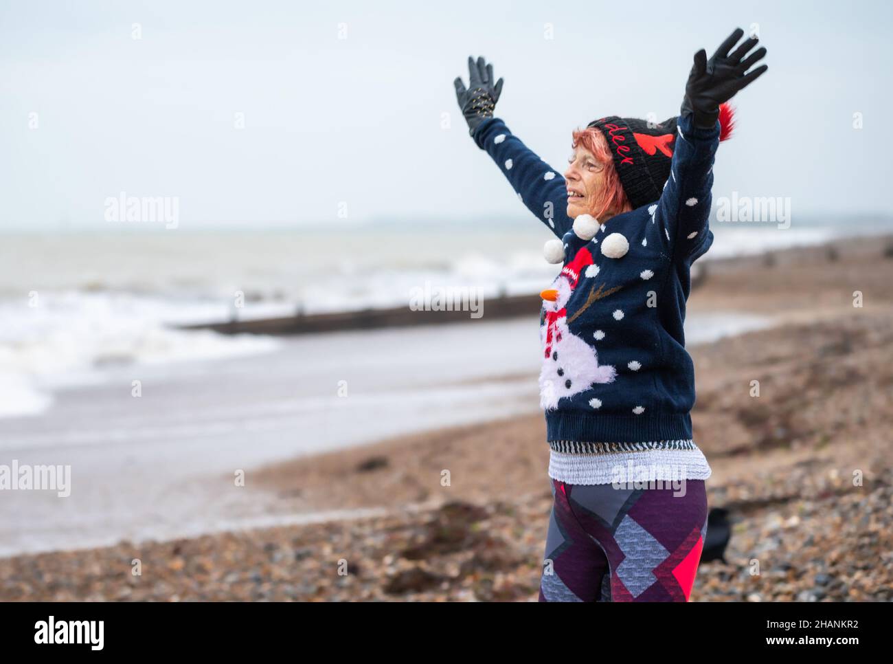 Ältere ältere Frau mit 80s Jahren an einem Strand, die im Winter trainiert. Sie trägt einen weihnachtlichen Pullover und Hut, während sie an der Südküste, Großbritannien, Sternsprünge macht Stockfoto