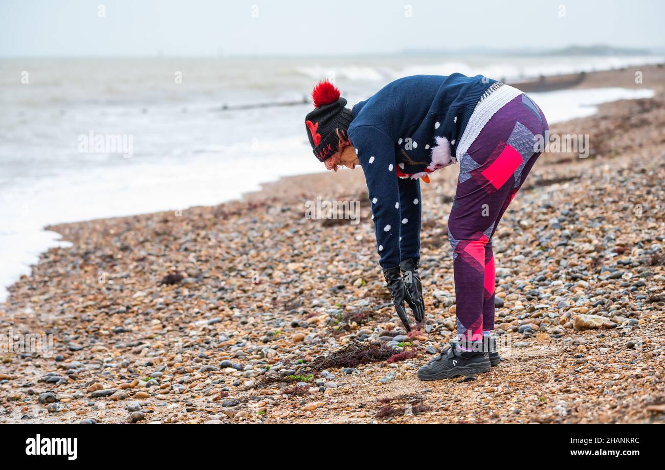 Ältere ältere Frau mit 80s Jahren an einem Strand am Meer, die im Winter trainiert. Die alte Dame trägt an der Südküste, Großbritannien, einen weihnachtlichen Pullover und Hut. Stockfoto