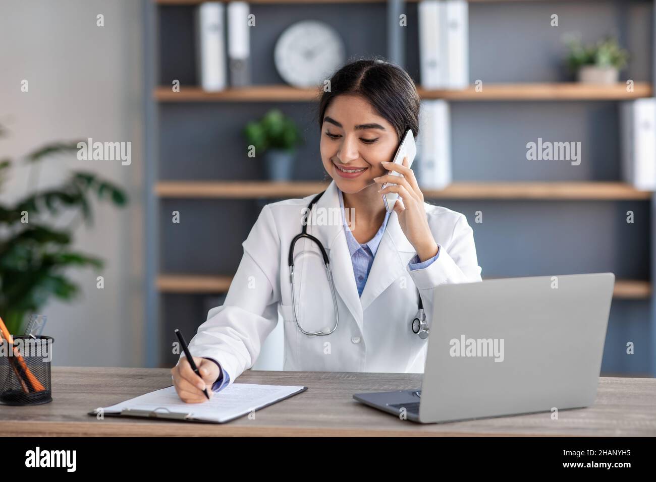 Lächelnd hübsche junge hindu-Ärztin im weißen Mantel mit Stethoskop telefoniert mit dem Patienten, arbeitet im Büro Stockfoto