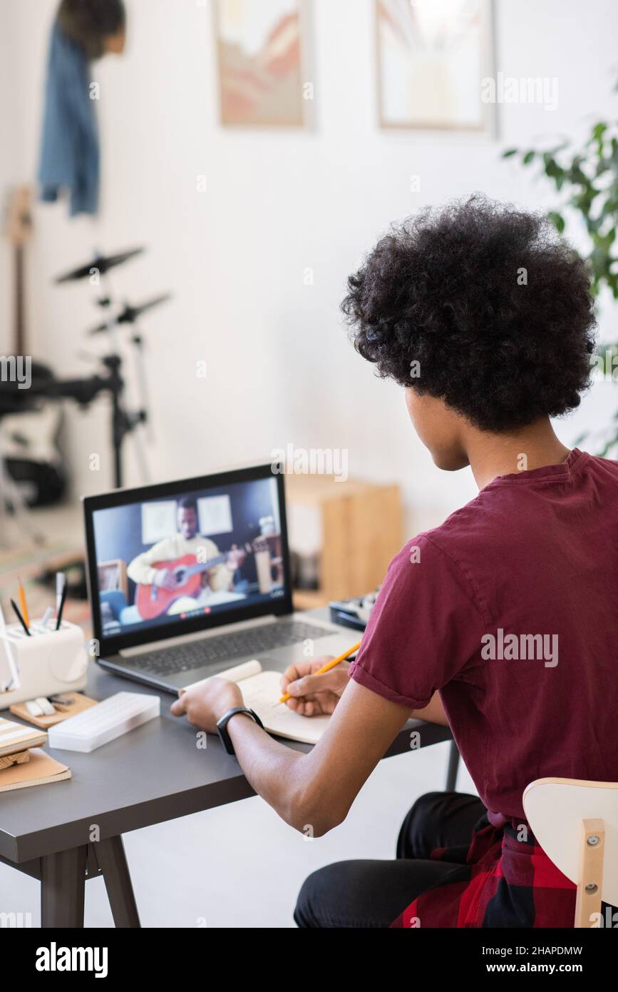 Biracial Kerl Notizen machen, während sitzen am Tisch und Blick auf Musiklehrer mit Gitarre auf Laptop-Bildschirm Stockfoto