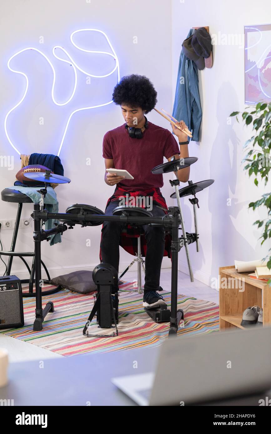 Moderner Biracial Youngster mit Drumkit sitzenden Drumsticks vor dem Laptop während des Trainings in seinem Schlafzimmer Stockfoto