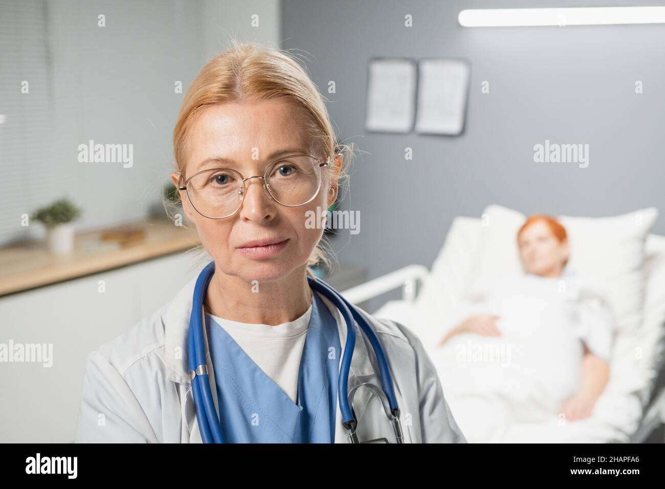 Porträt einer reifen Ärztin in einer Brille, die auf die Kamera schaut, während sie auf der Krankenstation steht und die Patientin im Hintergrund hat Stockfoto