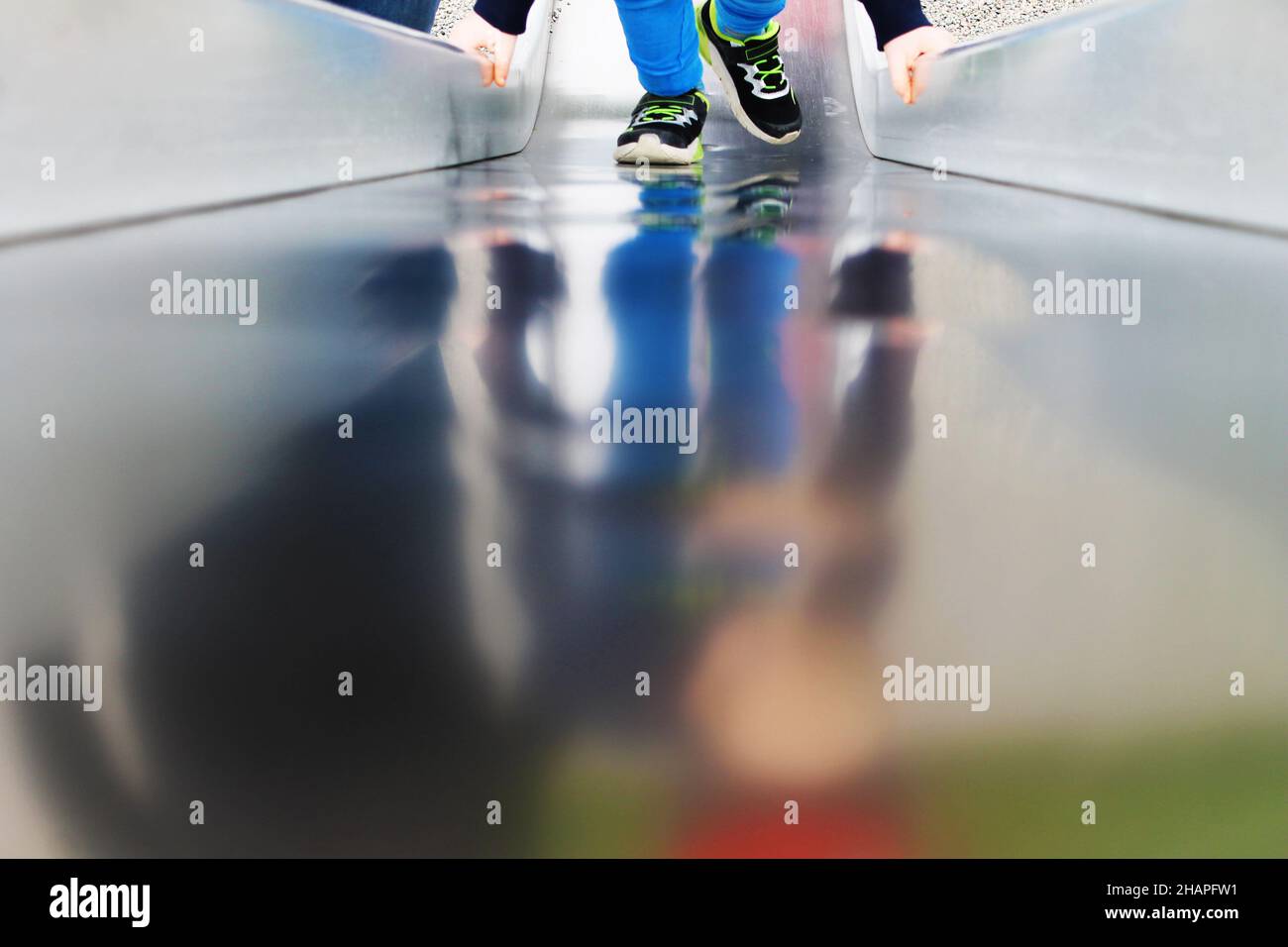Teilansicht eines Kindes auf einer Spielplatzrutsche, Spiegelung in Metalloberfläche. Stockfoto