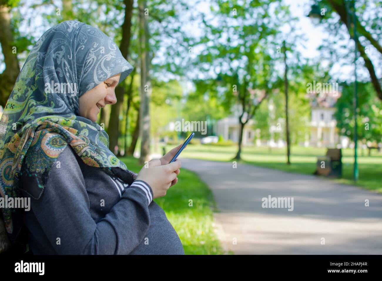 Profilansicht der muslimischen Schwangeren, die lächeln, während sie das Handy benutzen und allein im Park sitzen Stockfoto
