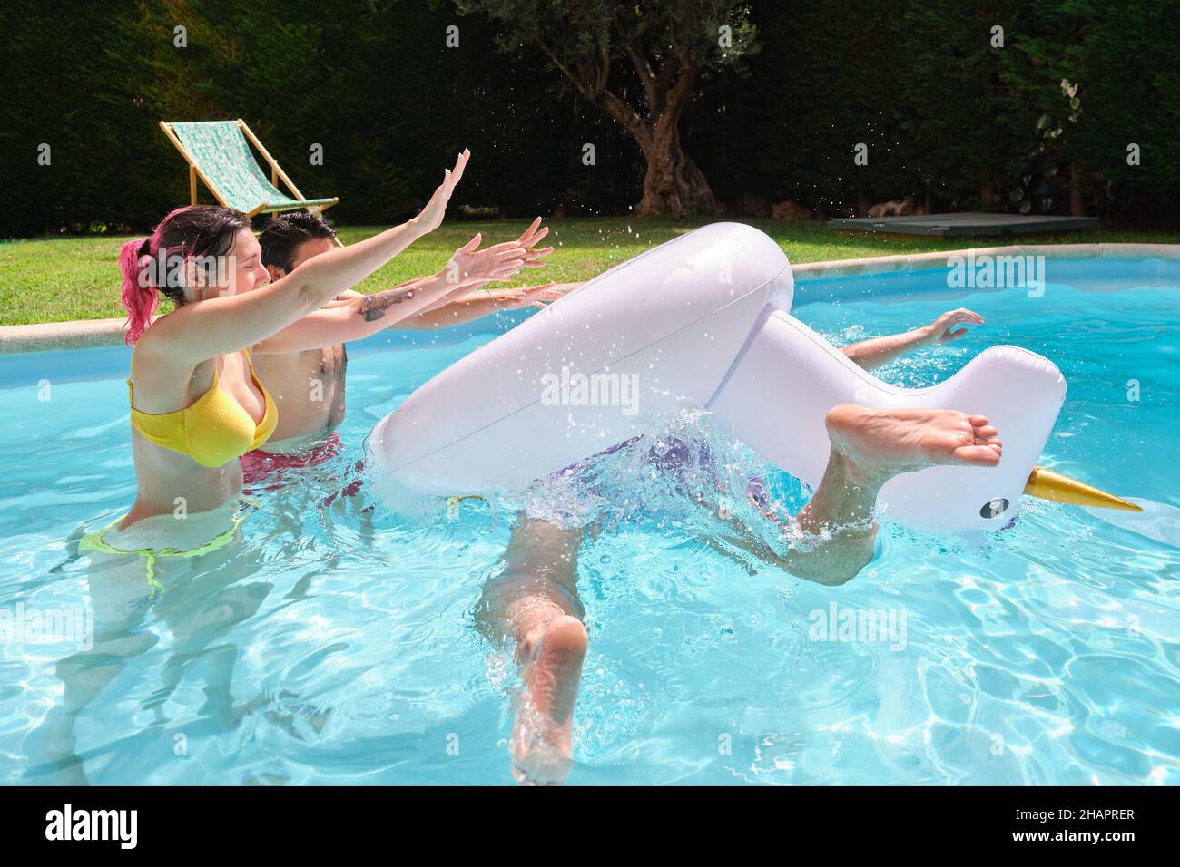 Zwei Freunde, die einen Mann aus einem Regenbogen-Einhorn werfen, schweben in den Pool. Stockfoto