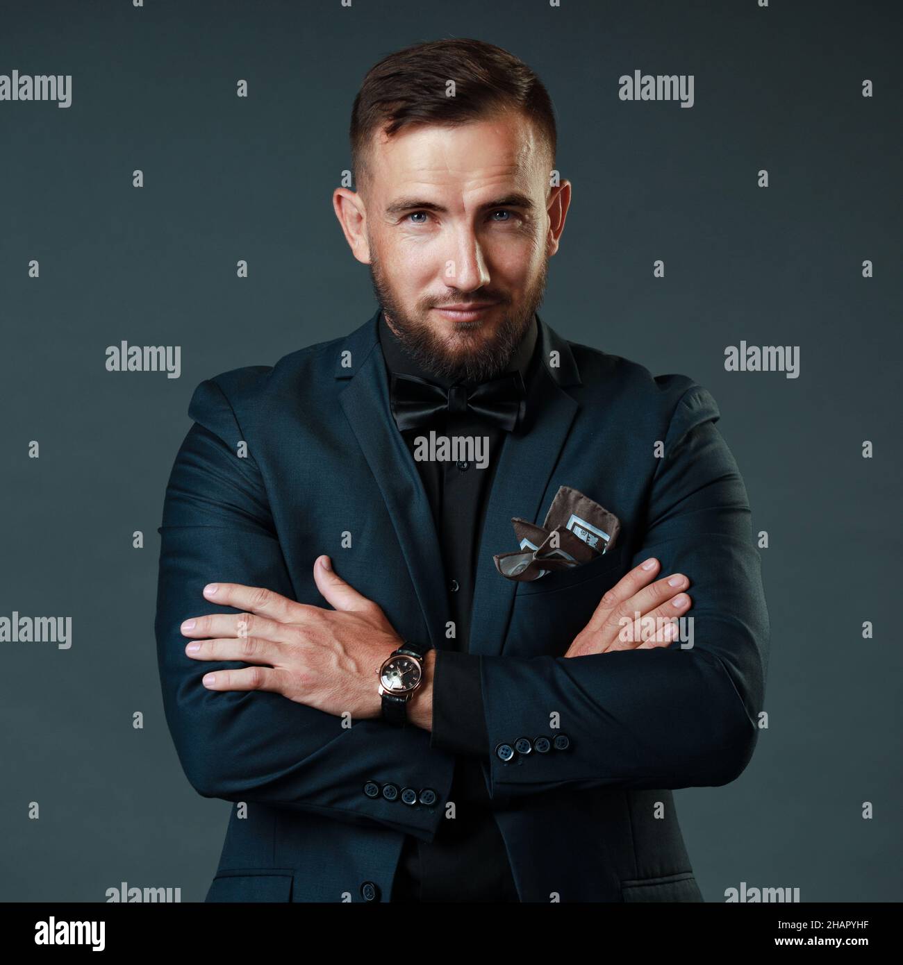 Attraktiver kaukasischer Mann in Jacke kreuzte seine Arme gegen seine Brust. Er sieht es mit einem Auge an. Studio-Porträt des Geschäftsmanns von 30 Jahren auf grauem Hintergrund. Übersinnlich oder verführerisch. Stockfoto