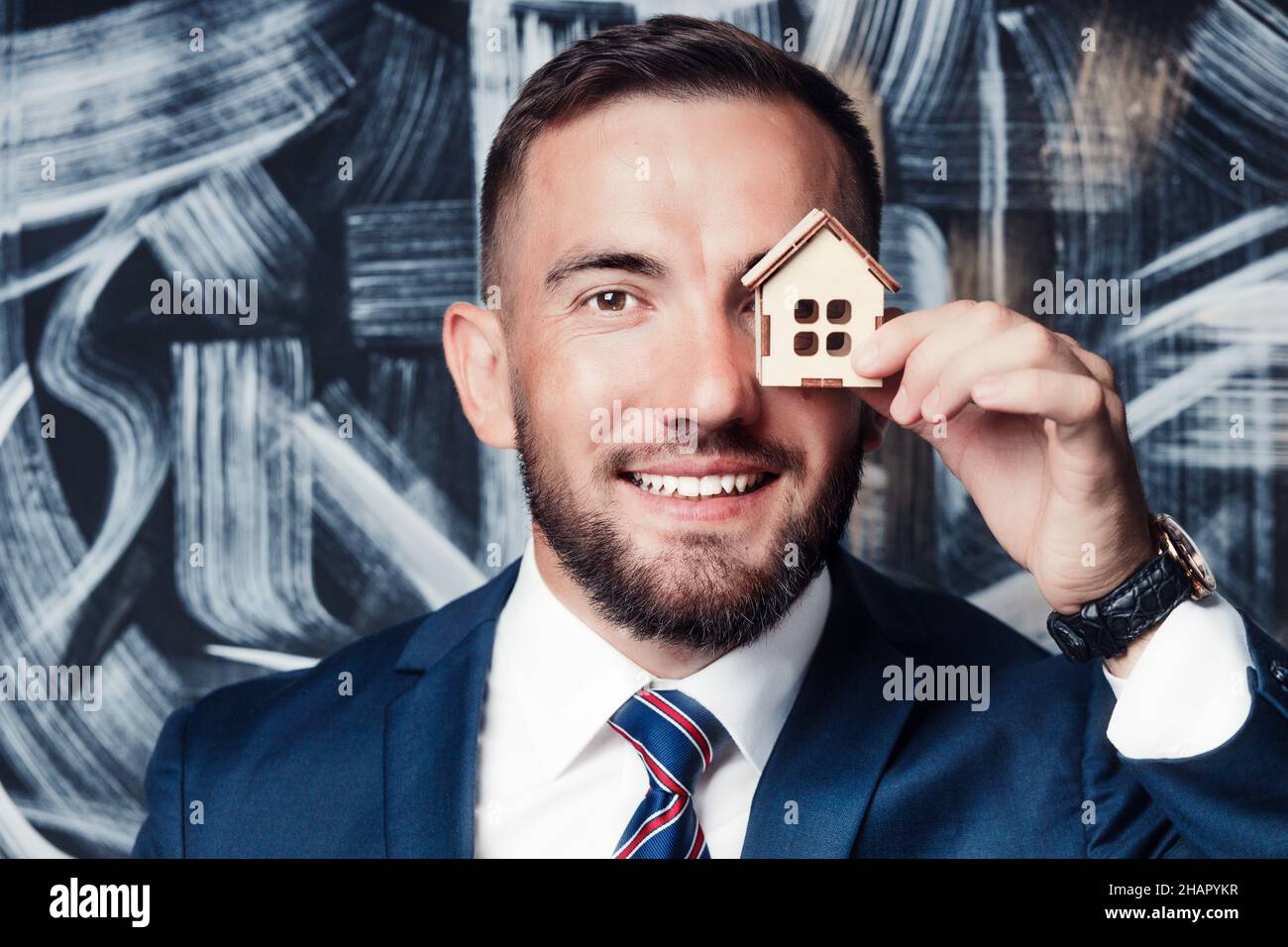 Nahaufnahme eines Immobilienmaklers oder Versicherers in Jacke und Krawatte. Der 25-35-jährige Kaukasusjunge hält ein Spielzeughaus in der Hand und lächelt. Hintergrund. Stockfoto