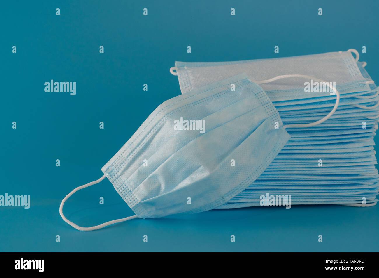 Es gibt viele neue medizinische chirurgische dreischichtige Masken, die hoch gestapelt sind und daneben liegt eine gebrauchte Maske auf blauem Hintergrund Stockfoto