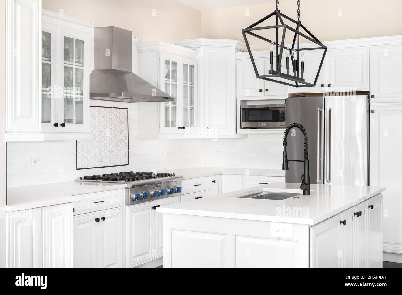 Eine schöne, moderne Küche mit weißen Schränken, Thermador-Geräten aus Edelstahl, einem schwarzen Kronleuchter über einer Insel und Marmorarbeitsplatten. Stockfoto