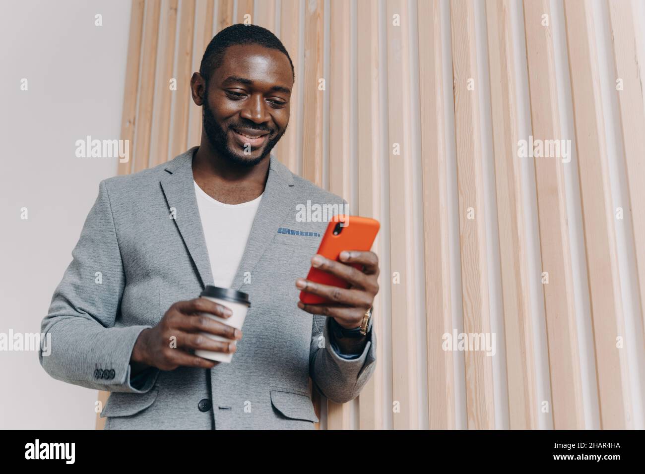 Kaffeepause bei der Arbeit genießen. Junger positiver afrikanischer Büroangestellter in Anzug mit Mobiltelefon Stockfoto