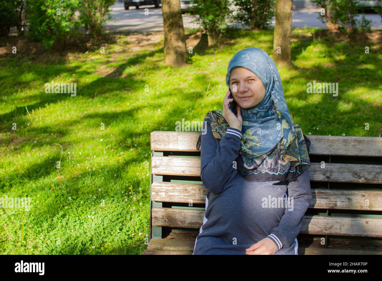 Junge muslimische Schwangere, die im Park sitzt und am Telefon telefoniert Stockfoto
